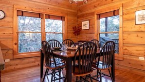 Una sala da pranzo in legno con un tavolo rotondo e sei sedie, ampie finestre con vista sugli alberi e un lampadario.