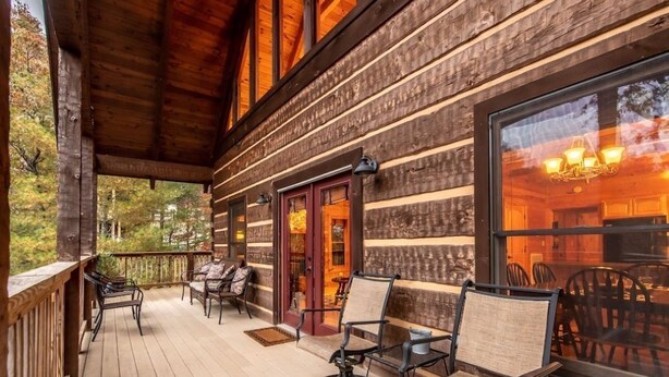 Una baita di legno con un portico, arredata con sedie e un tavolo, e un'ampia finestra che riflette l'illuminazione interna.