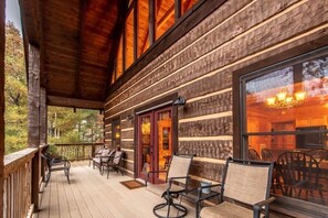 Une cabane en bois dotée d’un porche, aménagé avec des chaises et une table, et d’une grande fenêtre qui reflète la lumière intérieure.