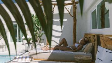 A person lying on a bed in a room with a pool, surrounded by plants and a pergola.