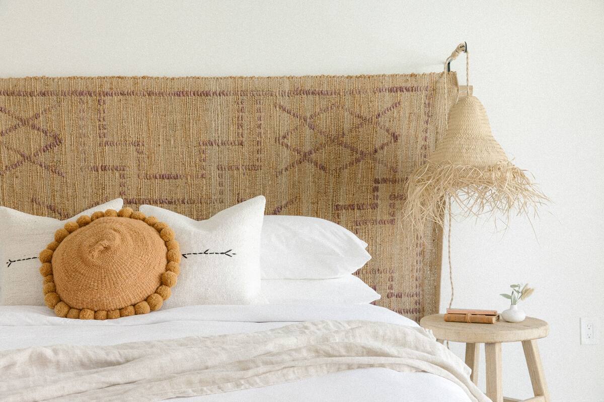 A neatly made bed with white linens, a woven headboard, and a round, textured pillow.