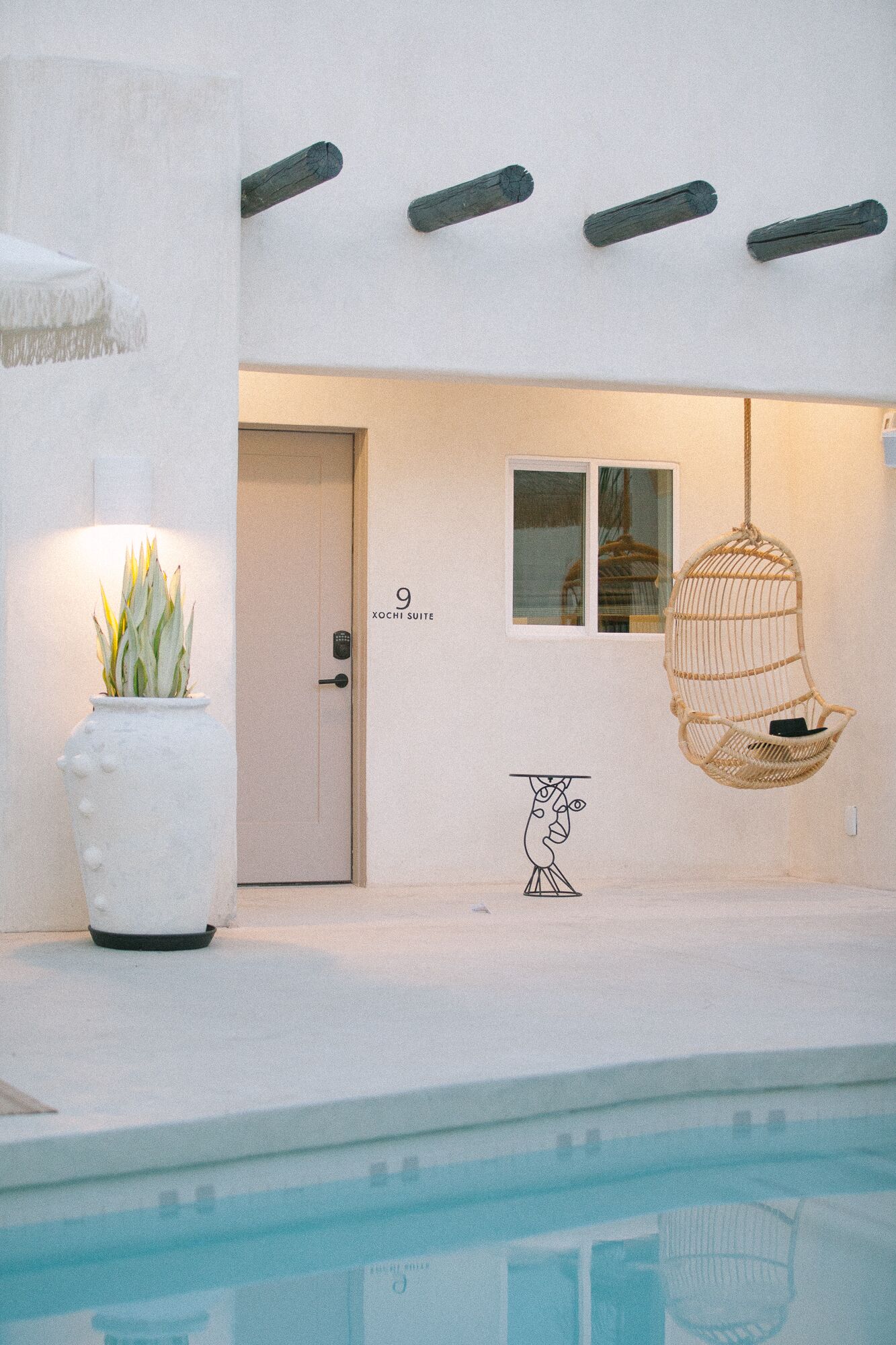 A modern building with a pool, a door labeled "Xochi Suite", a hanging chair, and a potted plant.