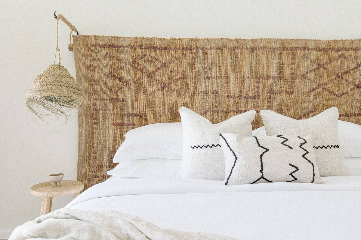 A bed with white bedding and patterned pillows, a woven headboard, and a small wooden nightstand with a bowl.