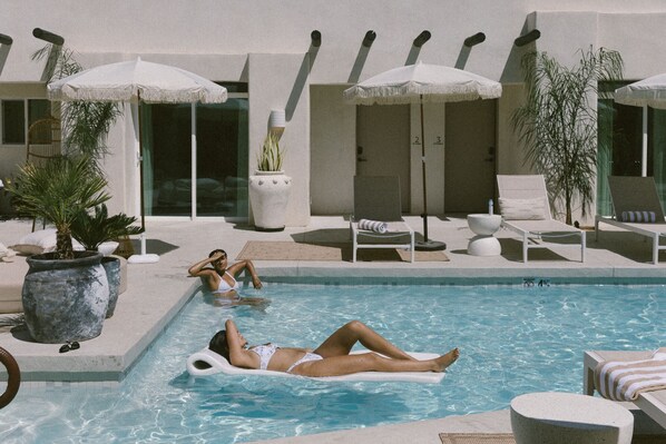 Un espace piscine avec des chaises longues et des parasols, une plante en pot et une personne se relaxant sur un flotteur gonflable.