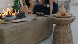 Two people sitting on a beige sofa outdoors with a fire, a wicker table with pottery, and a house in the background. - Yara Hotel (Palm Springs)