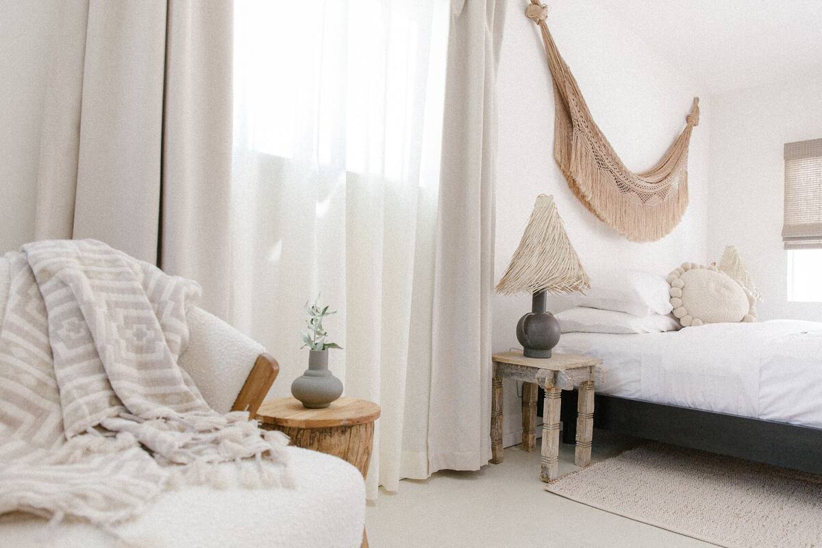 A bedroom with a bed, a chair, a nightstand with a lamp, and a macrame hanging.