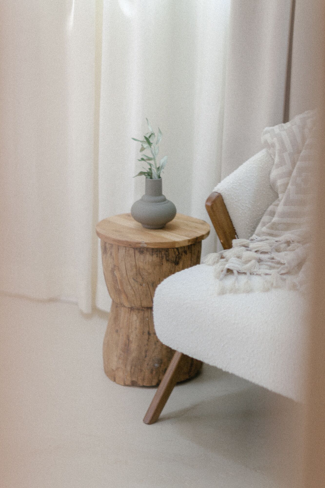 A room with a white sofa, a wooden side table with a vase of green plants, and a white curtain in the background.