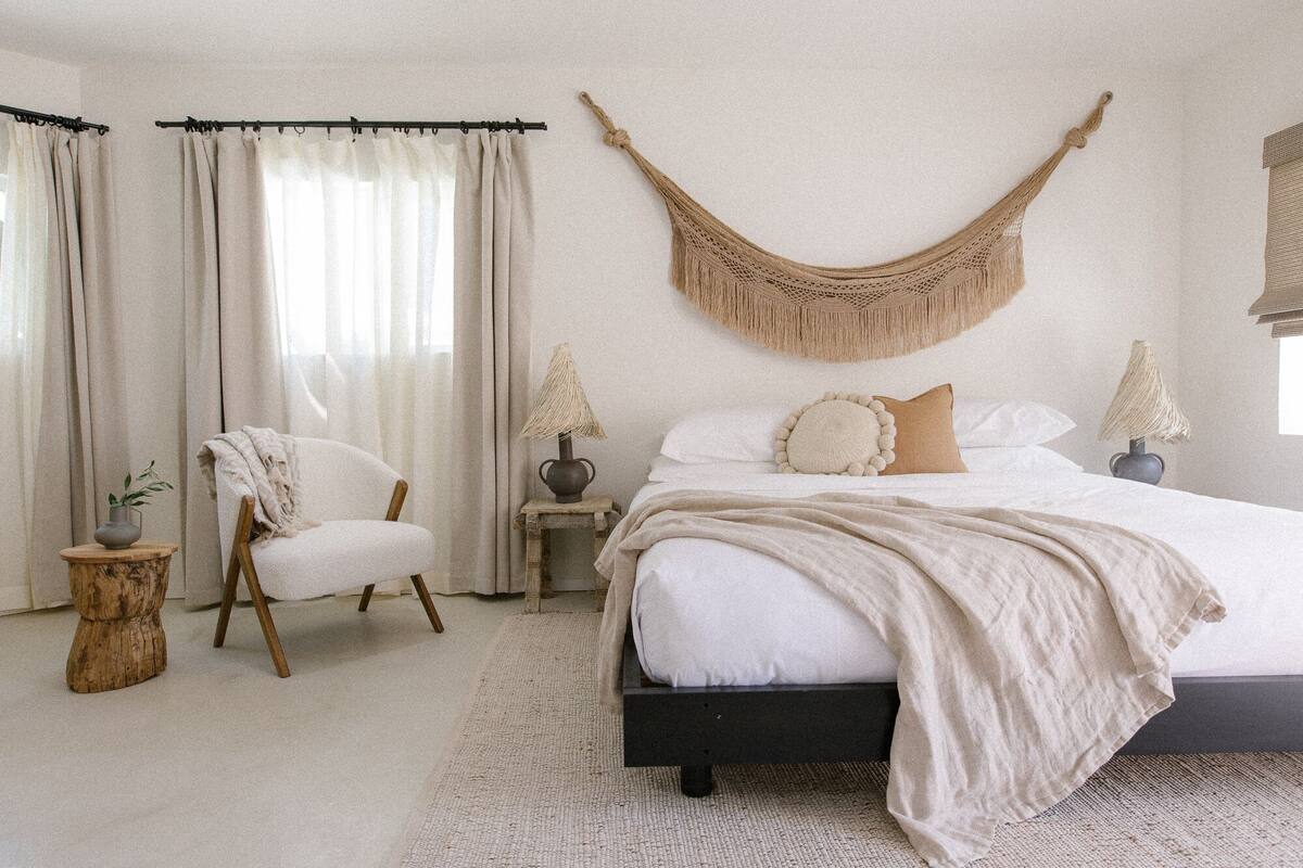 A bedroom with a bed, a chair, a macrame wall hanging, and a window with curtains.