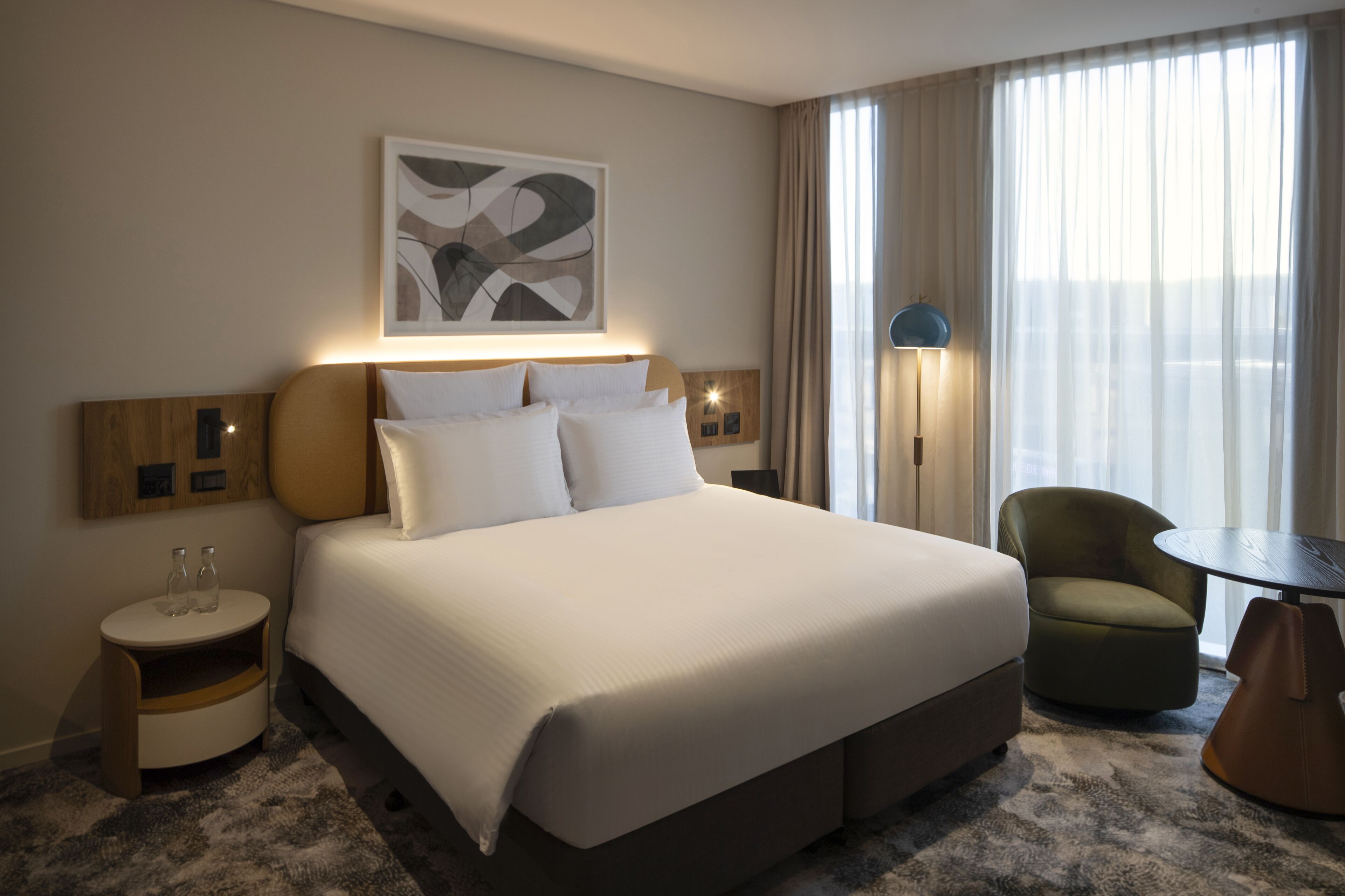 Habitación de hotel con una cama grande, una mesita de noche con vasos, una silla, una lámpara y un cuadro en la pared.
