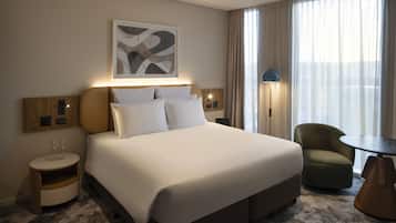 Habitación de hotel con una cama grande, una mesita de noche con vasos, una silla, una lámpara y un cuadro en la pared.