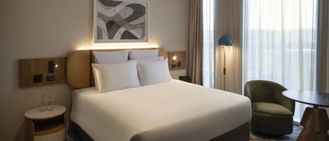 Habitación de hotel con una cama grande, una mesita de noche con vasos, una silla, una lámpara y un cuadro en la pared.