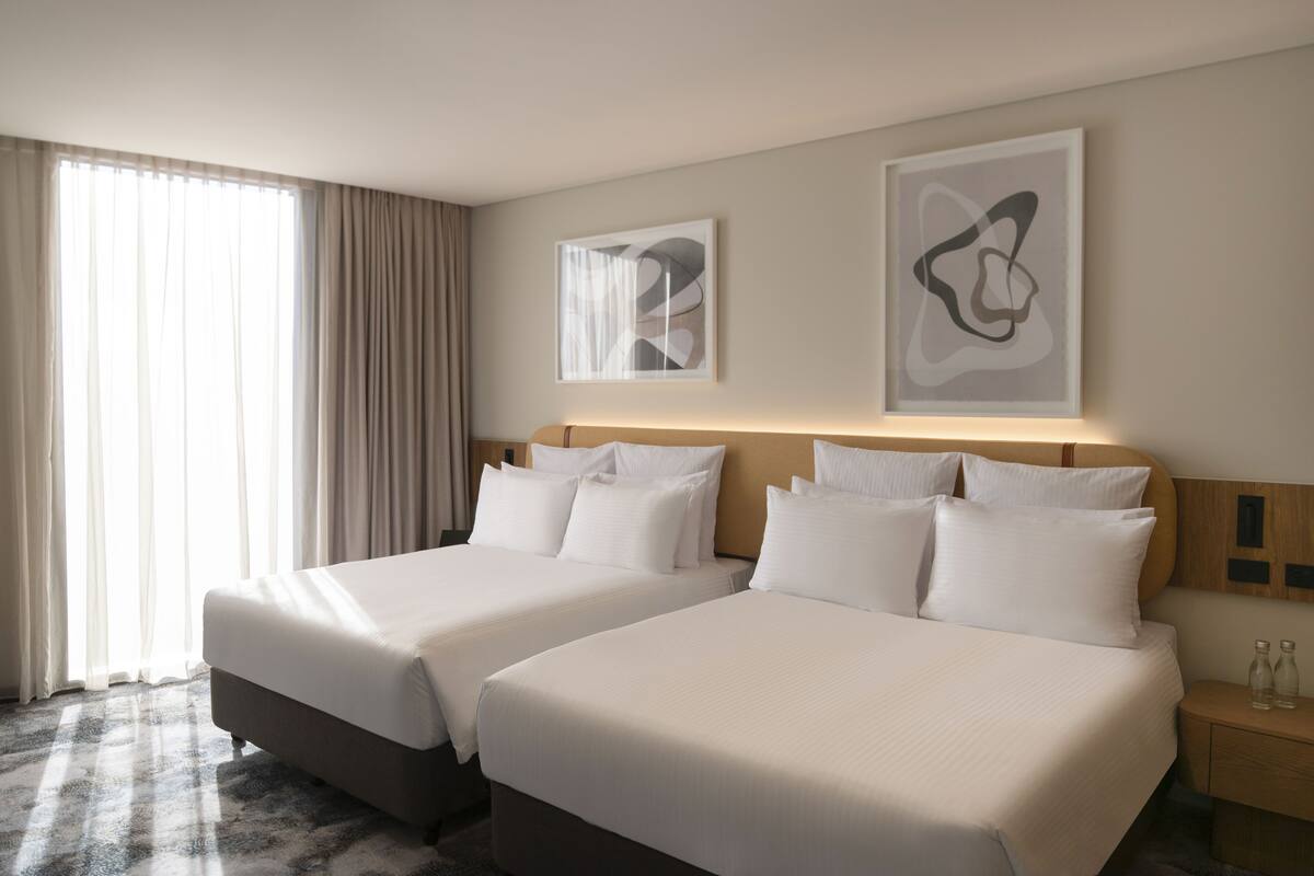 A hotel room with two beds, a large window with curtains, and two abstract art pieces on the wall.