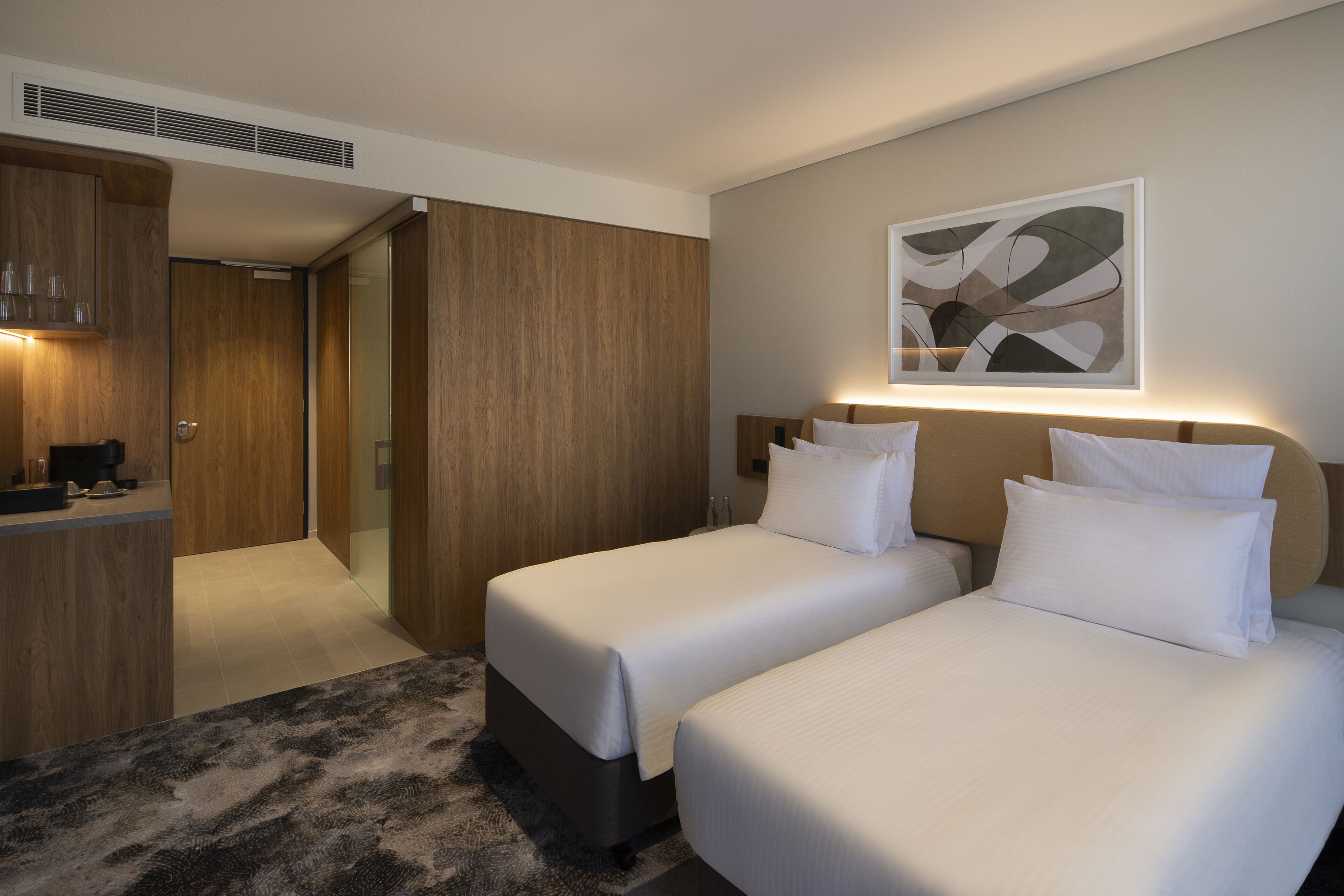 A hotel room with two beds, a wooden wardrobe, a kitchenette, and a modern abstract painting on the wall.