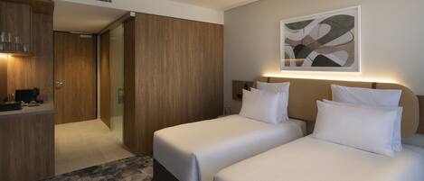 A hotel room with two beds, a wooden wardrobe, a kitchenette, and a modern abstract painting on the wall.