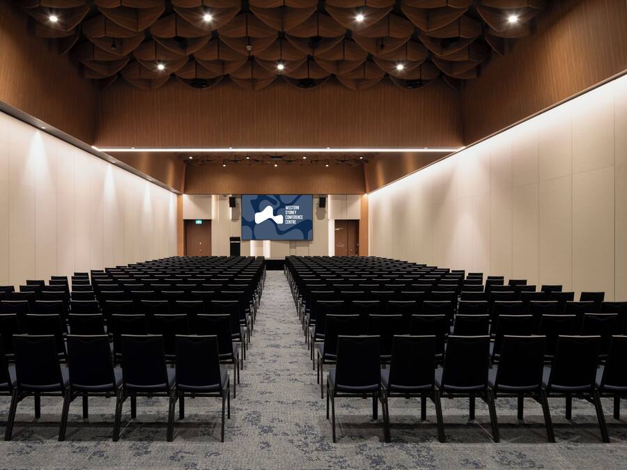 A conference room with rows of black chairs, a wooden ceiling, and a large screen at the front.