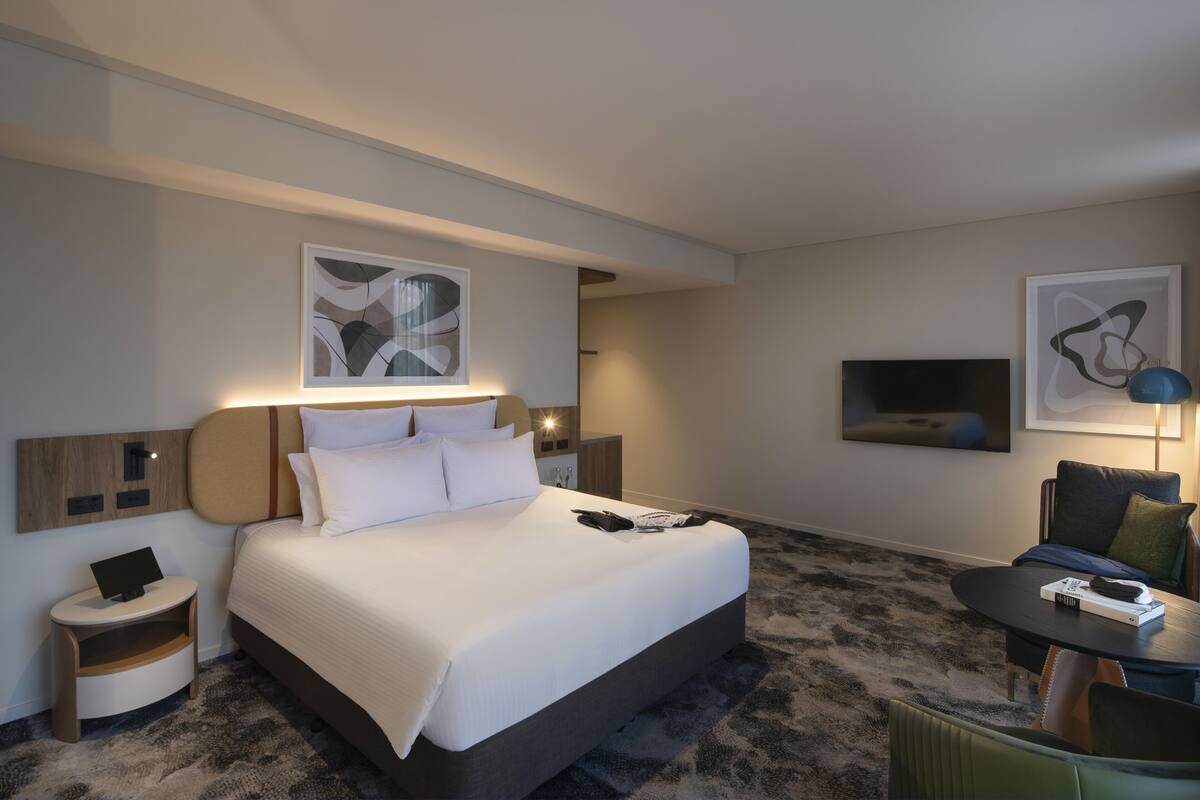 A modern hotel room with a large bed, a small side table, a TV mounted on the wall, and a seating area with a table and chairs.
