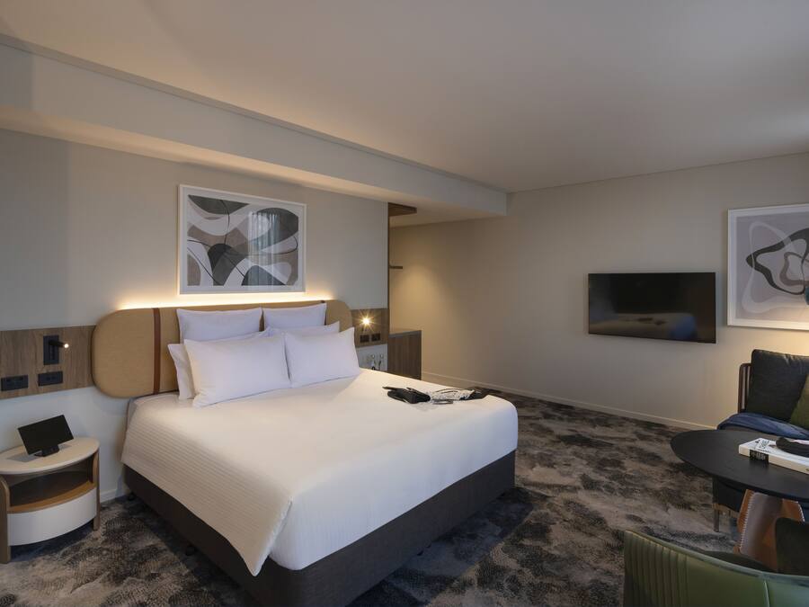 Una habitación de hotel moderna con una cama grande, una mesita auxiliar, un televisor montado en la pared y una zona de estar con una mesa y sillas.