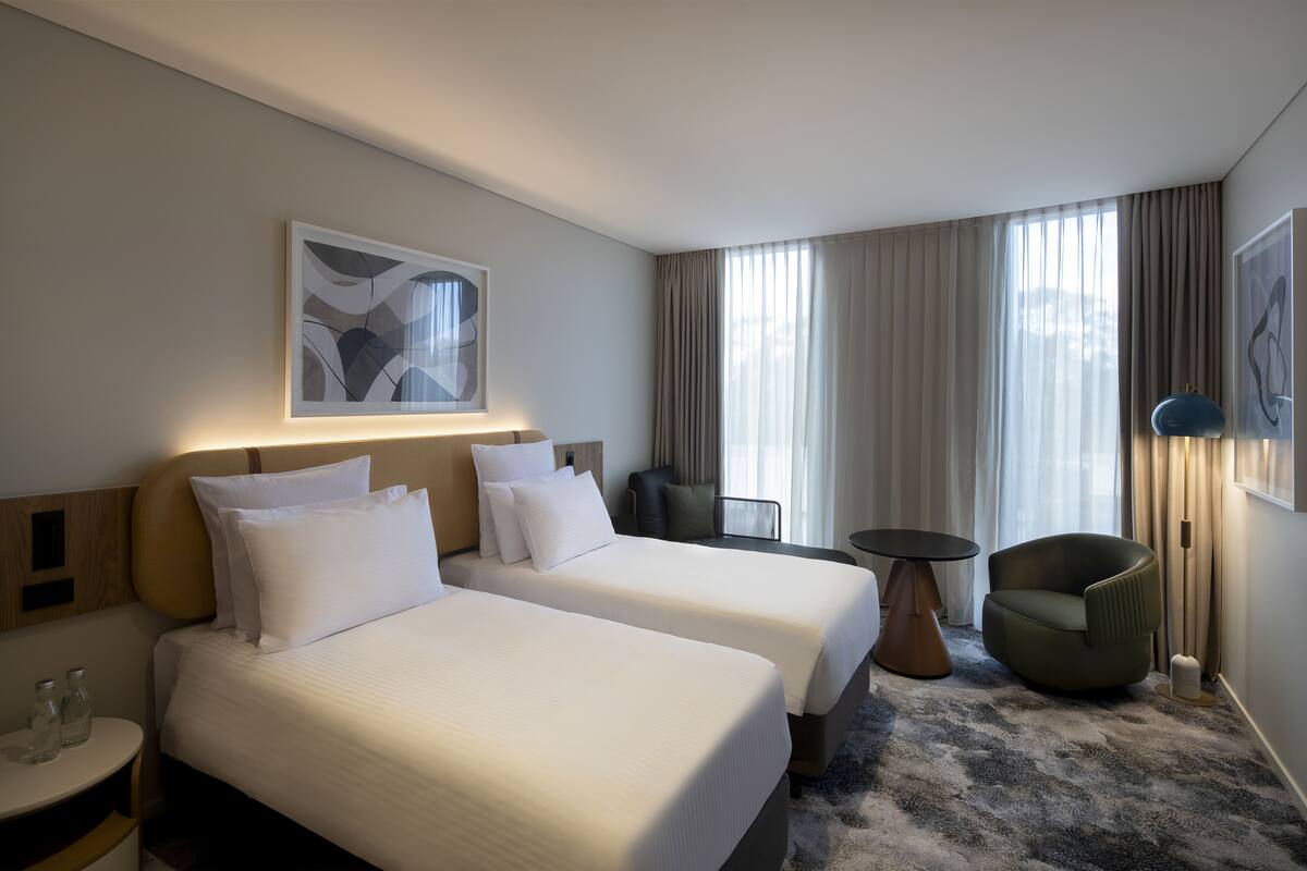 A hotel room with two beds, a small table, a chair, and a large window with curtains.