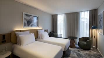 A hotel room with two beds, a small table, a chair, and a large window with curtains.