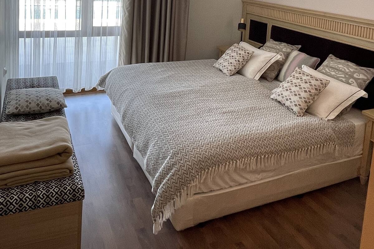 Een slaapkamer met een groot bed, een nachtkastje, een bankje en een raam met gordijnen.