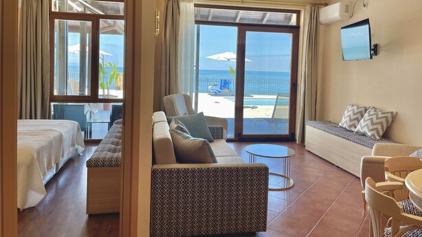 Une chambre d’hôtel avec un lit, un canapé, une petite table et un téléviseur. La chambre dispose d’une grande fenêtre donnant sur la plage et l’océan.