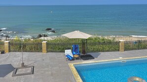 Une piscine avec vue sur l’océan, une plage et un espace de détente avec des chaises et un parasol.