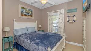 Ein Schlafzimmer mit einem Bett, zwei Nachttischen, einem Deckenventilator, einem Gemälde, einem Fenster mit Vorhängen, einer Rettungsboje als Dekoration und einem Fernseher.