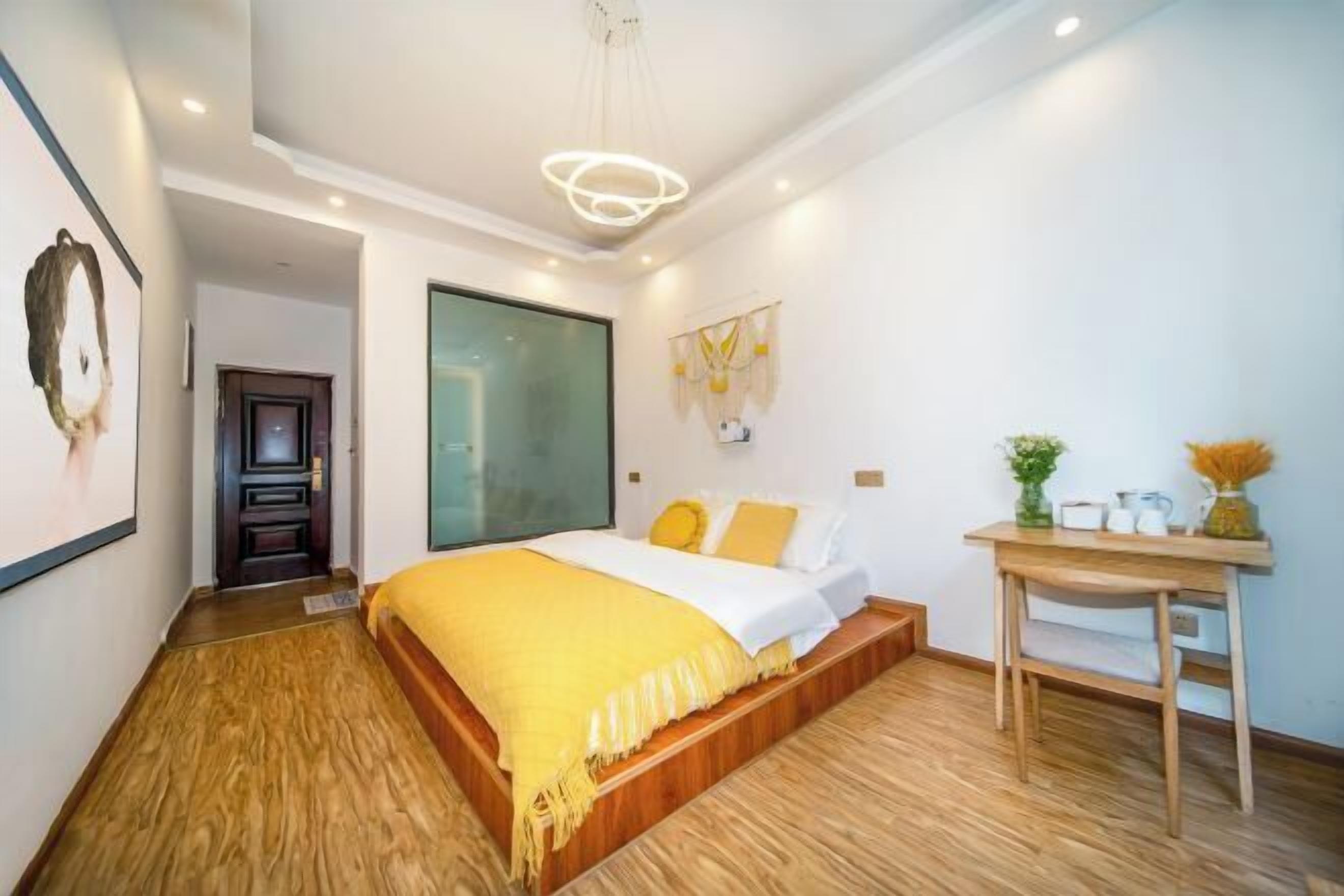 Un dormitorio moderno con piso de madera, una cama con un plaid amarillo, un escritorio con un florero y un ventanal grande.