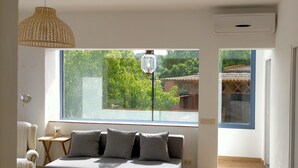Een slaapkamer met een groot raam, een bed met grijze kussens, een hanglamp en een wandmodel airconditioner.