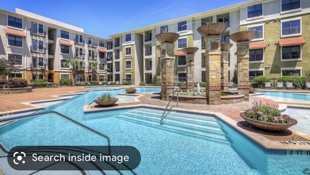 A multi-story hotel with a central swimming pool, stone pillars, and a clear blue sky.
