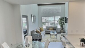 A modern living room with a glass dining table, white sofas, and a large window with blinds.