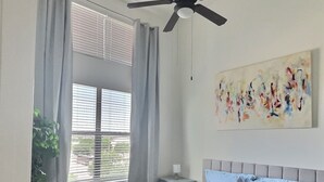 A bedroom with a bed, a ceiling fan, a window with curtains, a bedside table, and a painting on the wall.