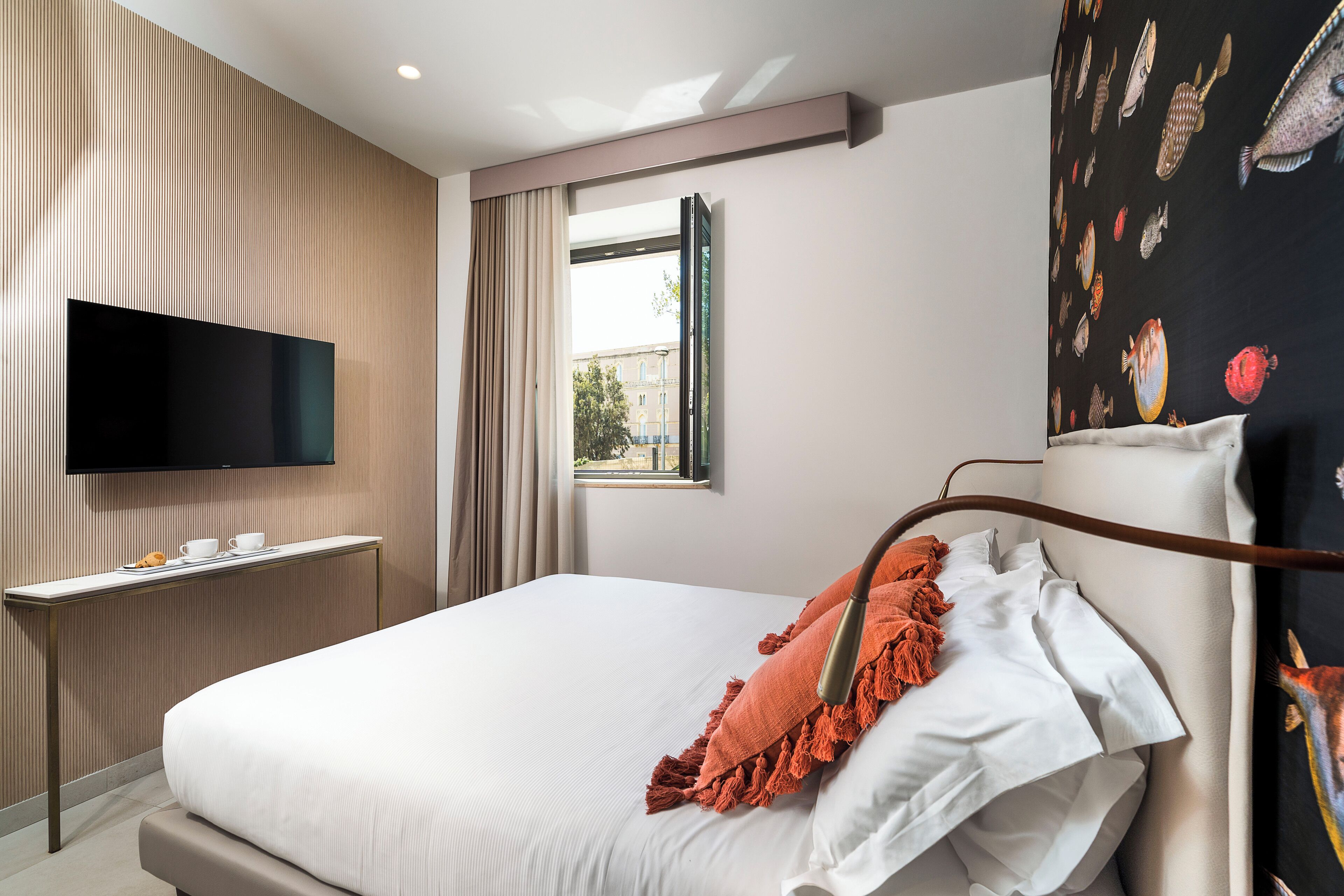 A modern hotel room with a large bed, a flat-screen TV mounted on the wall, a small table with a cup and saucer, and a window with curtains.