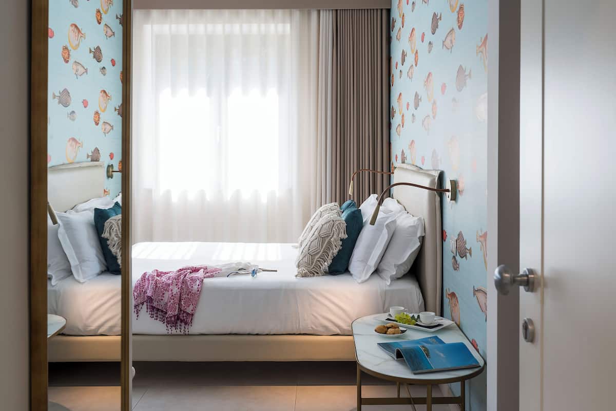 Quarto de hotel com uma cama, uma mesa de cabeceira com uma chávena e um livro, uma janela com cortinas e uma porta.