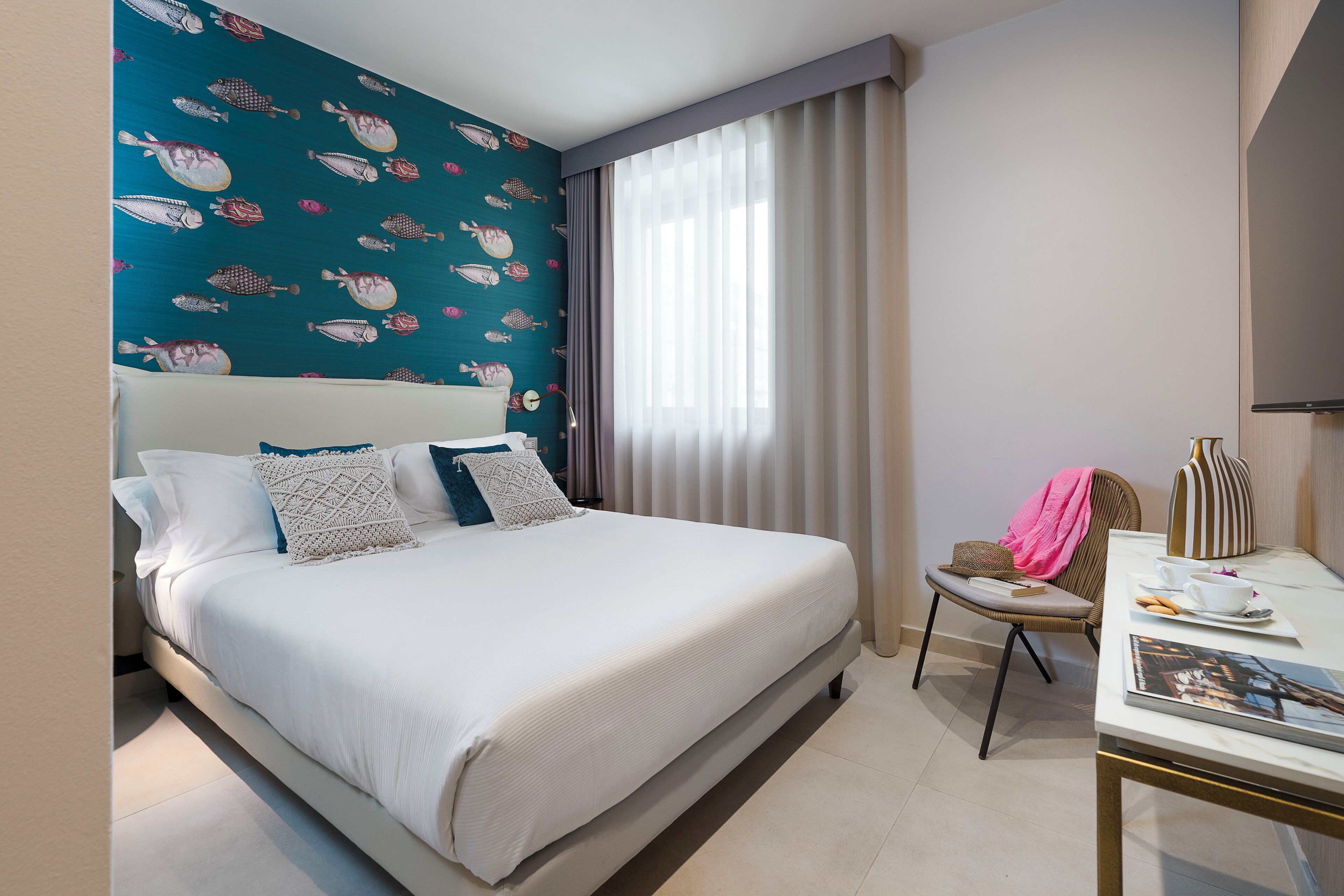 Een hotelkamer met een groot bed, een kleintje tafeltje met een tijdschrift, een stoel en een raam met gordijnen.