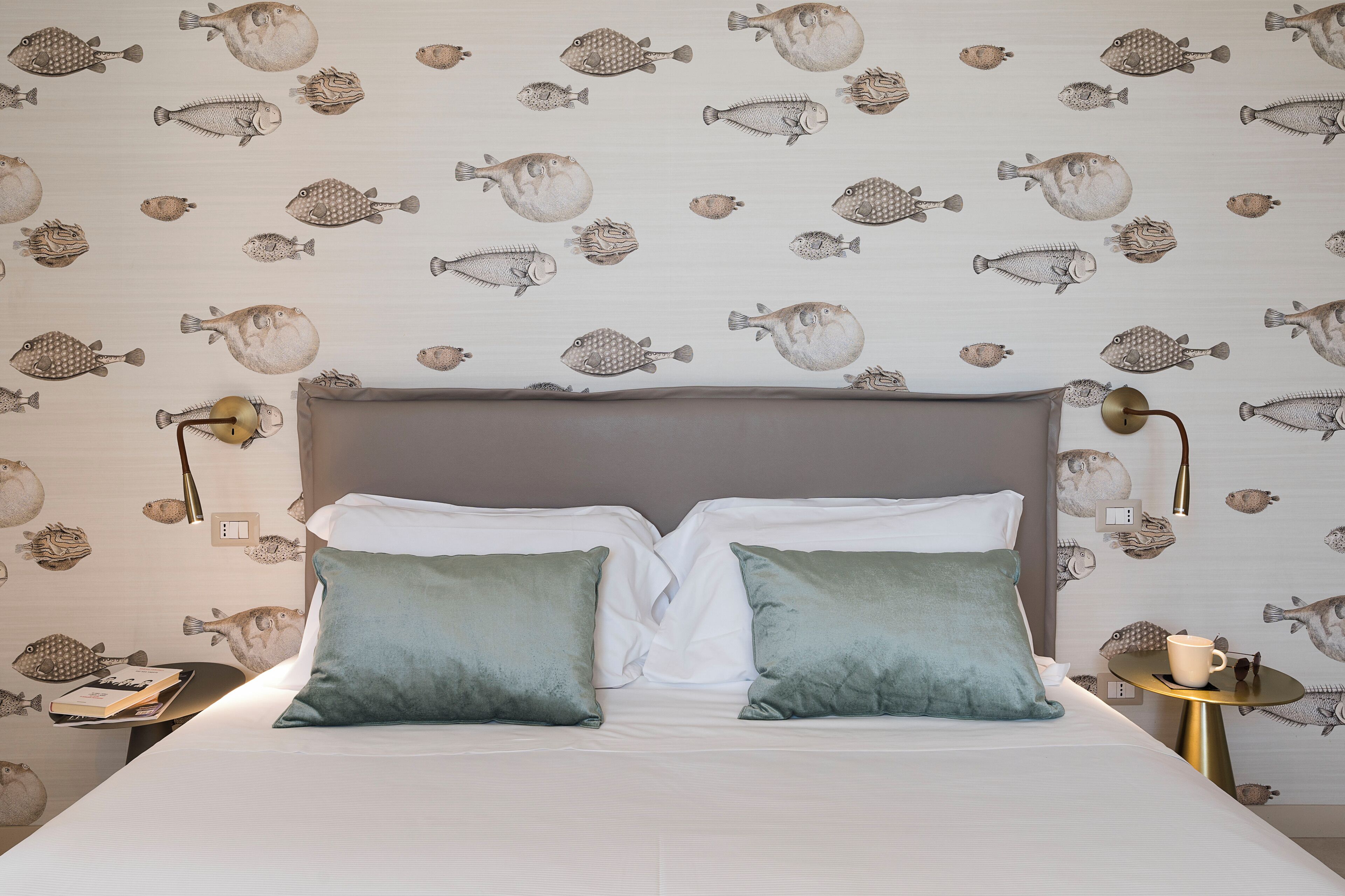 A hotel room with a bed, two pillows, a bedside table with a lamp, and a wall with fish wallpaper.