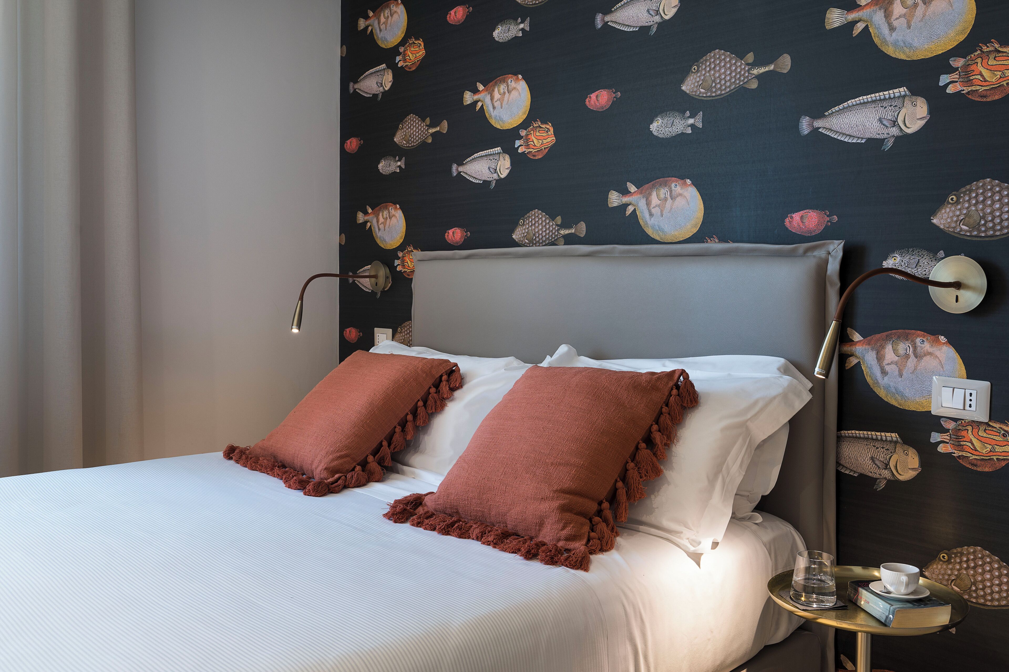 A neatly made bed with white linens and two brown pillows, a bedside table with a glass of water and a book, and a wall with a fish pattern.
