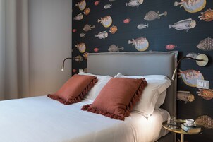 A neatly made bed with white linens and two brown pillows, a bedside table with a glass of water and a book, and a wall with a fish pattern.