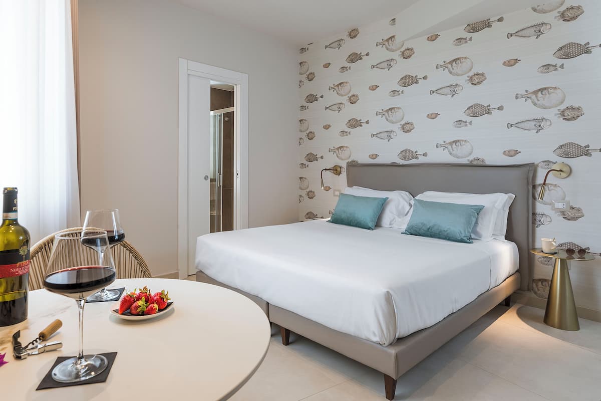 Quarto com cama, mesa de cabeceira com abajur, mesa de jantar com vinho e morangos, e porta que dá para outro cômodo.