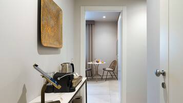 A modern hotel room with a minimalist design, featuring a desk with a laptop, a coffee machine, and a trash bin. There is a dining area with a table and chairs visible through an open door.