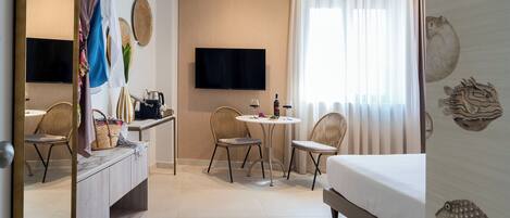 A hotel room with a bed, a TV, a dining table with chairs, a small kitchen area, and a window with curtains.