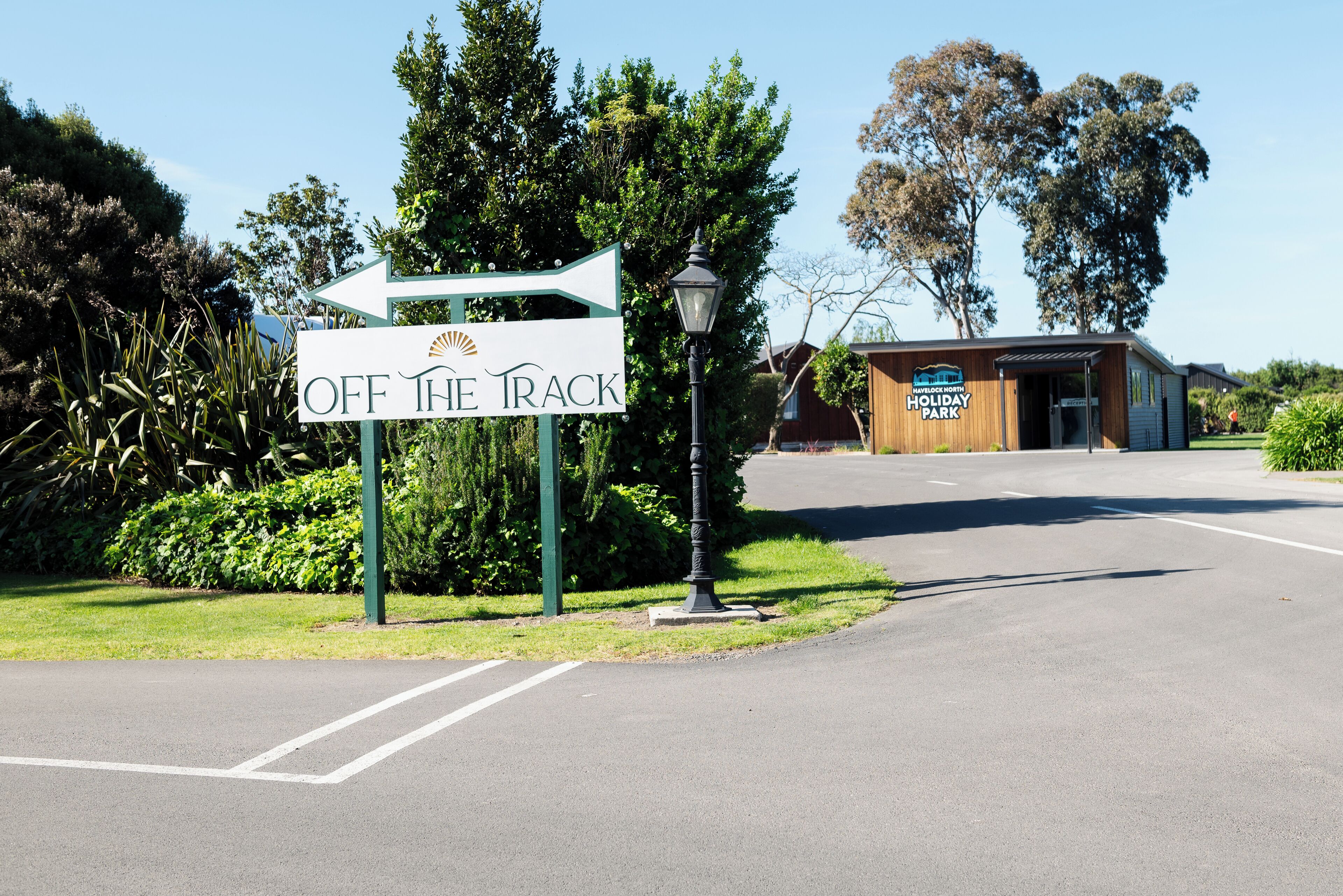 A sign for 'OFF THE TRACK' holiday park with a directional arrow, a lamp post, and a building with a reception area.