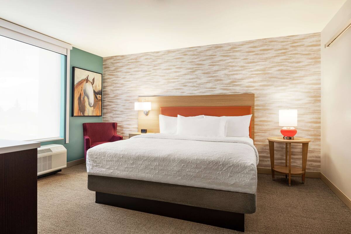A hotel room with a large bed, a desk, a chair, a lamp, and a painting on the wall.
