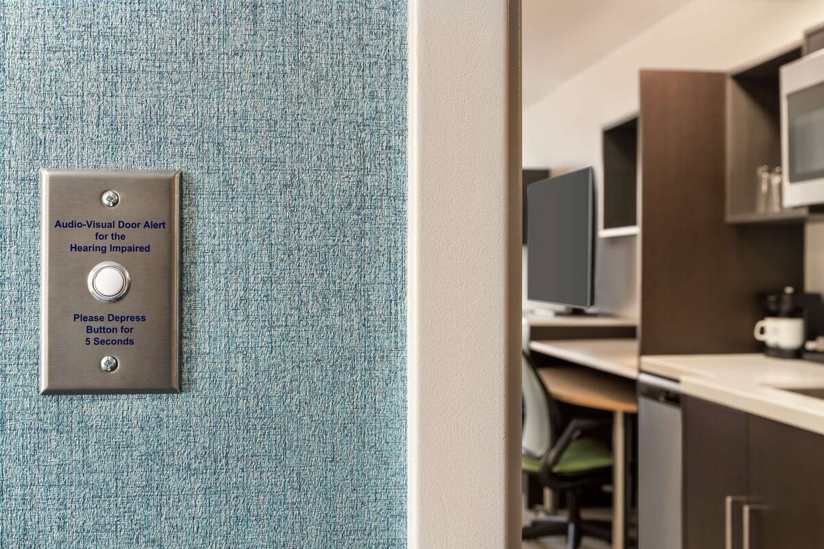 A door with an audio-visual alert system for the hearing impaired, a button to depress for 5 seconds, and a view into a modern kitchen area.