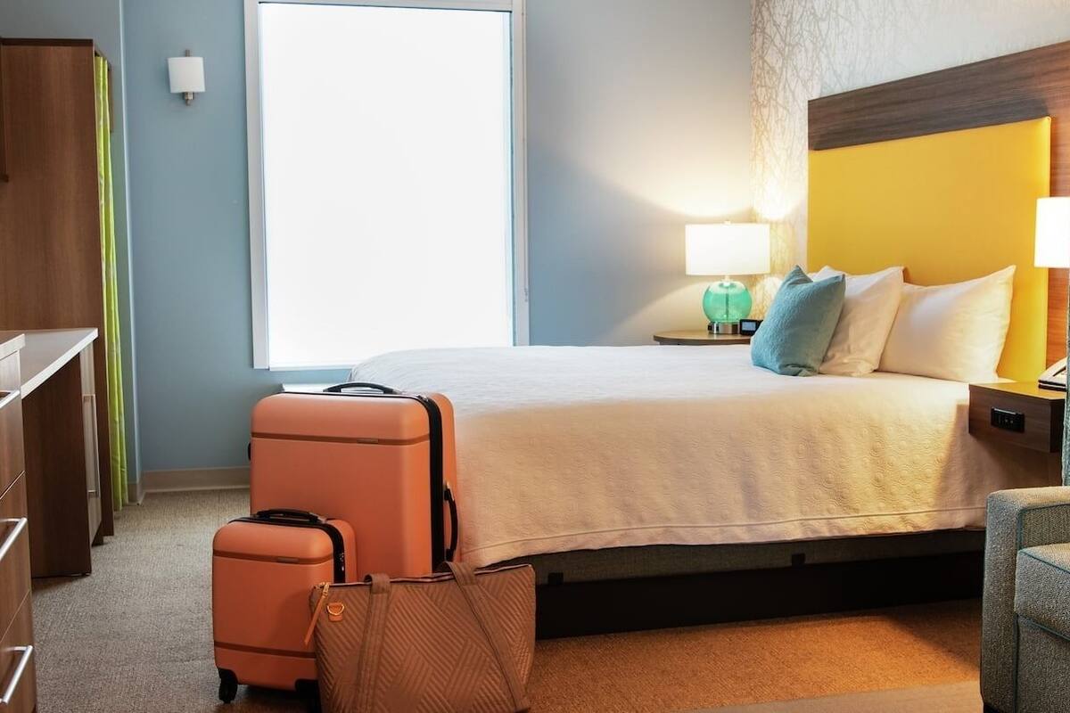 A hotel room with a bed, a desk, a chair, a suitcase, and a lamp.