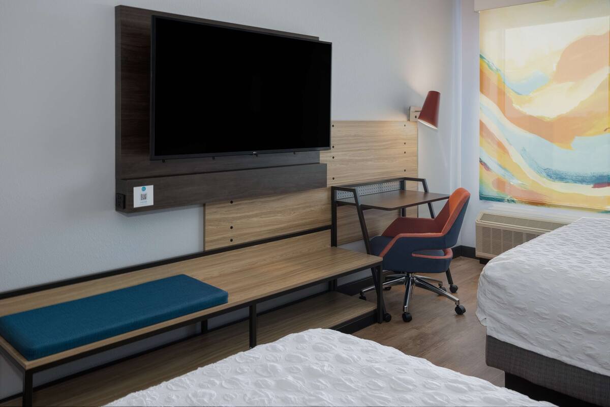 Une chambre d’hôtel équipée d’un téléviseur à écran plat fixé au mur, d’un bureau avec une chaise, d’un banc et de deux lits.