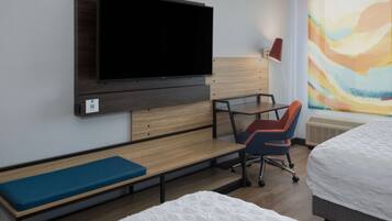 Een hotelkamer met een flatscreen televisie aan de muur, een bureau met een stoel, een bankje en twee bedden.