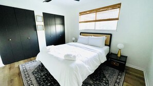 Un dormitorio con cama, ventilador de techo, ventana con persianas, mesita de noche con lámpara y armario oscuro.