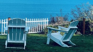 Duas cadeiras Adirondack voltadas para a água, com uma cerca branca e vista para montanhas ao longe.