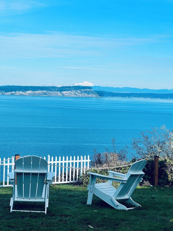 Two Adirondack chairs facing a body of water with a white picket fence and a view of distant mountains.